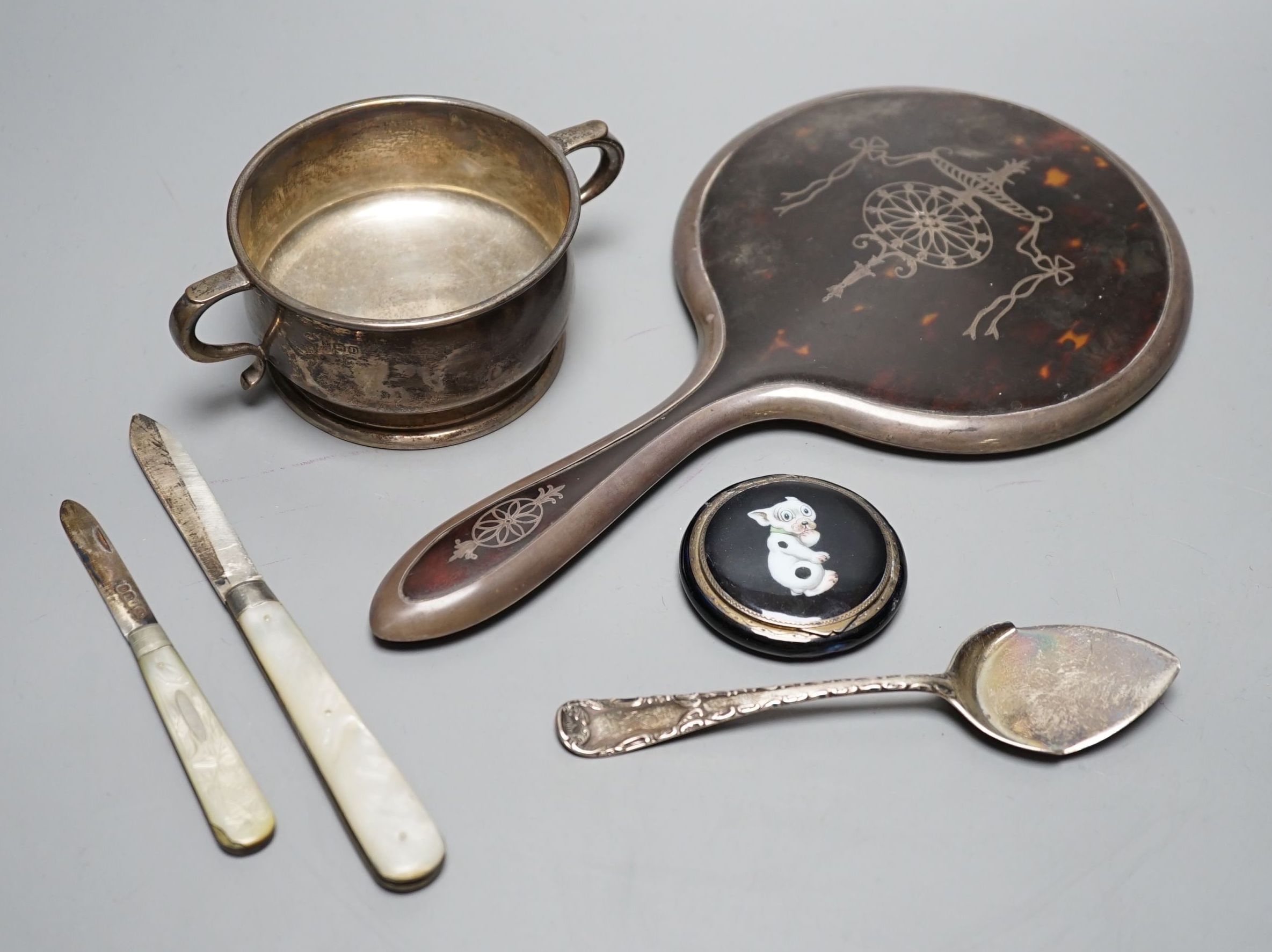 A silver two handled bowl, a silver mounted hand mirror, two silver and mother of pearl fruit knives, a silver preserve spoon and enamelled compact decorated with a dog.